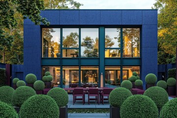 Denim blue modern villa surrounded by emerald green topiaries, with large windows and a maroon outdoor dining set