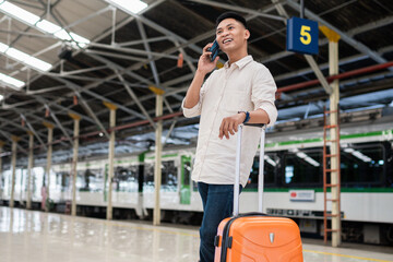 Cheerful traveler talks on a mobile phone at a train station platform. He stands with luggage, exuding excitement and anticipation. The setting suggests a journey or business trip