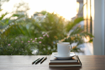 White coffee cup and notebook with lush green plant on wooden table under sunlight in the garden