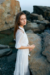 Poster - Woman in white dress standing on rocky shoreline with rocks in background, looking out at sea