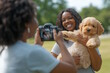 © MarGa - Smiling African American woman holding a golden doodle puppy while her friend takes a photo in the park, capturing a joyful moment. Concept of pet adoption and joyful moments in the community