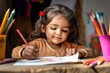 © Viktor - Young Indian girl in brown dress drawing with red crayon on white paper. Vibrant colors, red, yellow, green, blue. Desk with art supplies, pink, yellow, blue cups. Gray wall, window in background.