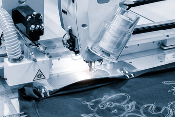 Poster - A sewing machine is in use, with a piece of fabric being sewn. The machine is blue and white, and the fabric is blue. The machine is in a factory setting, and the image conveys a sense of productivity