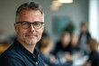 © pham - A professional man with glasses smiles at the camera while participating in a team discussion in a contemporary workspace