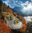 © den-belitsky - Aerial view of winding mountain road at sunset in autumn. Alps, Italy. Curved alpine road, snowy rocks, trees, red leaves, lake, sky with sunbeams. Colorful. Top view of road on mountain pass in fall