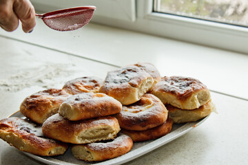 buns in a plate full of freshly baked cheesecakes a female hand sprinkled with powdered sugar on the kitchen table. cooking baking. stage of preparation of baking rolls. breakfast closeup copy space