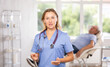 © JackF - Confident polite professional female doctor in blue scrub standing in medical office and making inviting gesture