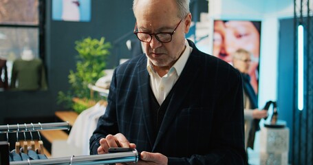 Canvas Print - Trendy senior man shopping for new fashion collection at retail store, visiting modern clothing shop to check formal or casual items. Rich customer uses phone to look for products by code. Camera B.