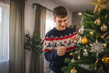 Young man decorate a Christmas tree with festive ornaments at home