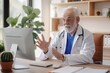 © miss irine - Old male doctor works in telemedicine. Sits at desk, wearing white coat, uses computer for online consultations. Gestures with hand, holds pen, potted plant adds plants to room. Natural enters