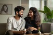 © miss irine - Young Indian couple sitting on gray couch in living room. Man and woman holding coffee cups, relaxed and happy. Gray walls and carpet create calm atmosphere. Window view of tree outside.