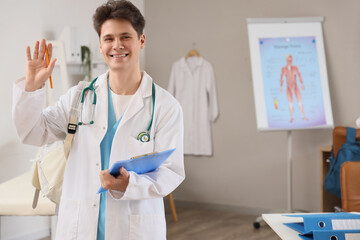 Wall Mural - Male medical intern with backpack and clipboard waving hand in clinic