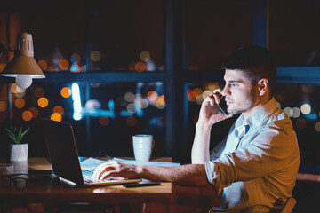 Poster - Overnight Work. Businessman At Laptop Talking On Phone Sitting Near Panoramic Window With Night City View In Office. Empty Space