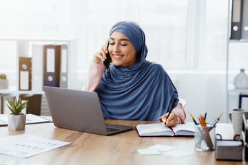 Poster - Business Lifestyle. Portrait Of Busy Middle Eastern Businesswoman At Workplace In Office, Talking On Cellphone, Using Laptop And Taking Notes