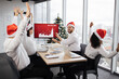 © sofiko14 - Successful team of multiethnic business people in Santa hats raising hands and screaming with happiness while working together in decorated with Christmas tree office in business center.