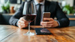 © D.e.a.r - A faceless businessperson in suit is holding smartphone while sitting at wooden table with glass of red wine. scene conveys sense of sophistication and modernity