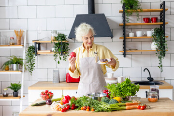 Wall Mural - happy beautiful elderly gray haired senior woman cook in cozy kitchen with fresh organic vegetables, smiling elderly woman makes choice between sweet and harmful unhealthy donut and healthy ripe apple