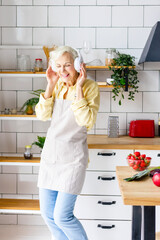 Wall Mural - happy beautiful elderly gray haired senior woman in headphones cook in cozy kitchen with fresh organic vegetables, having fun and singing