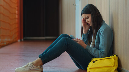 Sad upset failure stress pensive thinking girl female woman student pupil education studying sitting floor in school university college corridor using mobile phone smartphone online app social media