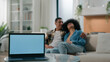 © Yuliia - Laptop with empty copy space gray blue screen advertise ads template foreground African American happy family talking couple man woman together at home couch talk conversation mortgage loan insurance