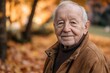 © ChaoticMind - A senior man in a brown jacket enjoys the crisp autumn air in a wooded area, with a gentle smile and vibrant orange leaves creating a peaceful atmosphere.