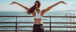 © Sascha - Slim sporty sportswoman standing on a pier with her arms outstretched looking out at the ocean enjoying the beautiful view and the calming sound of the waves