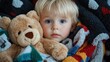 © familymedia - A toddler looks curiously at the camera, wrapped in a plush and colorful blanket. The child holds a teddy bear, representing innocence and warmth.