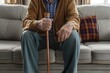 © Vadym - Elderly man sits on gray couch wearing brown sweater, blue plaid pants. Holds blue cane in right hand, purple, orange plaid pillow in left hand. Natural illuminates scene through white curtains.