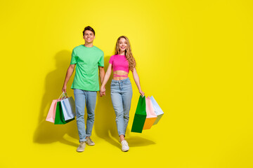 Poster - Full size photo of two nice young people hold hands store bags walk empty space isolated on bright yellow color background