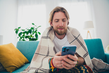 Photo of nice young man sit sofa cover blanket hold device wear stylish pajama modern light cozy interior flat indoors