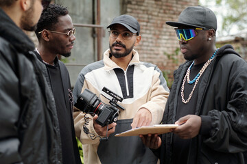 Poster - Group of men engaging in a conversation on set about film project while holding camera and clipboard. Diverse crew showing film equipment during planning phase without looking at camera