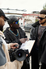 Leinwandbilder - Group of people engaging in project discussion while holding documents and camera outside. Urban backdrop and casual attire highlight collaborative and creative environment