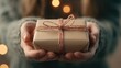© svastix - Hands offer a neatly wrapped gift with twine, set before a softly blurred background of warm, glowing lights, evoking generosity and holiday spirit.