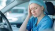 © supattra - Tired nurse woman in blue scrubs sitting behind the wheel of her car, she is holding her head with her hand and appears to be feeling fatigued by pain or fear.