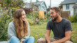 © Robert Kneschke - Happy young woman talking with male friend outdoors in a garden setting, sharing cheerful conversation with nature and homes in the background, showcasing a moment of friendship and relaxation.