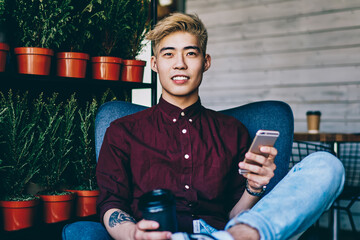 Positive chinese hipster blogger holding coffee to go in hand while resting in comfortable chair in modern cafe with loft interior, cheerful asian young man using smartphone device for communication