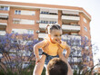 © BeatrizHerrera - A father lifts his smiling daughter above his head in a residential area. The scene captures their playful interaction against a backdrop of blooming trees and an apartment building.