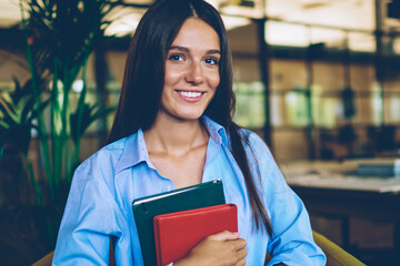 Portrait of happy cheerful hipster girl looking at camera before literature circle while holding new best sellers in hand and smiling, positive clever brunette female student posing with notepads