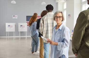 Wall Mural - Happy american voter registering at polling station on election day holding ballot paper. Smiling woman standing in a queue among citizens of USA at vote center and looking cheerful at camera.