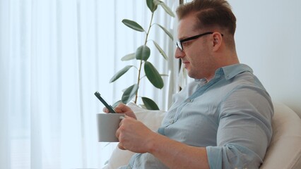 Middle age man sit on sofa with coffee cup in room using phone search data for occupation, raising children to be good adult or entertainment. Businessman use technology for convenience. Divergence.