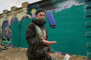 Guy in respirator, beanie cap and hoodie throwing blue can with paint while standing against green wall of building