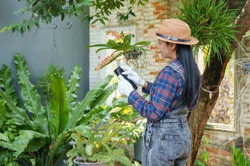 Asian woman using a tablet while tending to hanging plants in a home garden. Wearing gloves, plaid shirt, and hat, focused on organizing and managing plants in a fresh and green outdoor environment.