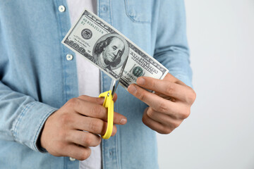 Budgeting. Man cutting dollar banknote with scissors on light grey background, closeup