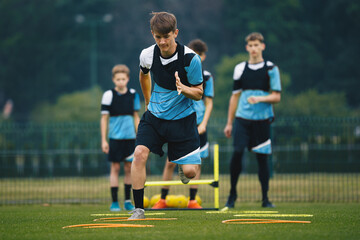 Wall Mural - Teenagers on soccer training camp. Boys practice football with young coaches. Junior-level athletes improving soccer skills on outdoor training. The player jumping over hurdles and ladder-skipping