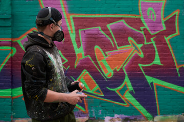 Side view of young man in casual attire and protective respirator holding cans with graffiti paints and looking at artwork on brick wall