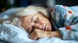 © svastix - An elderly woman rests quietly on a comfortable bed, enveloped in a cozy, patterned blanket, showcasing a tranquil and serene moment of peace in a domestic setting.
