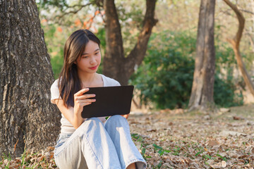 Young asian women writing business data and journey diary on digital tablet while sitting on the grass under tree in the nature park to working outside and relaxation with journey travel lifestyle