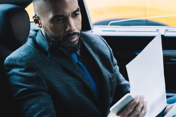 Dark skinned male strategist checking information for create productive plan for company development working on way to office building, business passenger in automobile reading contract details