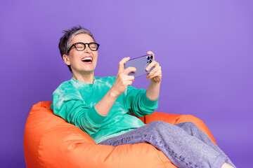 Poster - Photo portrait of pretty retired female sit beanbag laugh play mobile game wear trendy green outfit isolated on violet color background