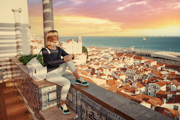 Wall Mural - Traveling by Portugal. Young traveling woman enjoying old town Lisbon view, red tiled roofs, ancient architecture and river on sunset